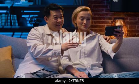 Heureux excité multiracial couple multiethnique marié à divers hommes asiatiques et femme caucasienne parlant en ligne conférence par appel vidéo avec application de téléphone mobile Banque D'Images