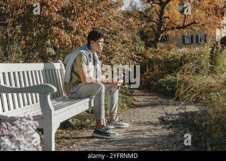 Un adolescent est assis sur un banc dans le parc boit du café dans une tasse thermo et regarde dans un téléphone. Portrait de beau gars joyeux assis sur le banc fres Banque D'Images