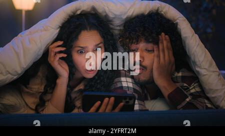 Couple multiracial homme et femme à la maison de nuit en utilisant le téléphone mobile choqué par les nouvelles en ligne choquer émerveillez-vous sur les réseaux sociaux de smartphone le soir Banque D'Images