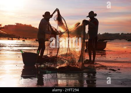 Pêcheurs vietnamiens démantelant les filets de pêche au soleil couchant. Mui ne, Vietnam Banque D'Images