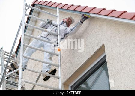 Maler schleift die alte Farbe auf den Holzteilen, dem Dachüberstand an einem Haus ab. Er steht auf einem mobilen Gerüst, Vorarbeiten zum Malern des Dachüberstands, modèle libéré für redaktionelle Nutzung, Dachüberstand streichen *** peintre ponçant la vieille peinture sur les parties en bois, le surplomb du toit sur une maison il se tient sur un échafaudage mobile, travaux préparatoires pour peindre le surplomb du toit, modèle libéré pour usage éditorial, peindre le surplomb du toit 20241105-DSC 8760 Banque D'Images