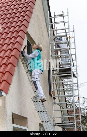 Malerin kratzt mit einer Drahtbürste die alte Farbe auf denHolzteilen, dem Dachüberstand an einem Haus ab. SIE steht auf einer Leiter, Vorarbeiten zum Malern des Dachüberstands, modèle libéré für redaktionelle Nutzung, Dachüberstand streichen *** le peintre utilise une brosse métallique pour gratter la vieille peinture sur les parties en bois, le surplomb du toit sur une maison elle se tient sur une échelle, travaux préparatoires pour peindre le surplomb du toit, modèle libéré pour usage éditorial, peindre le surplomb du toit 20241105-DSC 8775 Banque D'Images