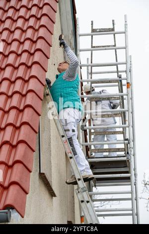 Malerin kratzt mit einer Drahtbürste die alte Farbe auf denHolzteilen, dem Dachüberstand an einem Haus ab. SIE steht auf einer Leiter, Vorarbeiten zum Malern des Dachüberstands, modèle libéré für redaktionelle Nutzung, Dachüberstand streichen *** le peintre utilise une brosse métallique pour gratter la vieille peinture sur les parties en bois, le surplomb du toit sur une maison elle se tient sur une échelle, travaux préparatoires pour peindre le surplomb du toit, modèle libéré pour usage éditorial, peindre le surplomb du toit 20241105-DSC 8778 Banque D'Images