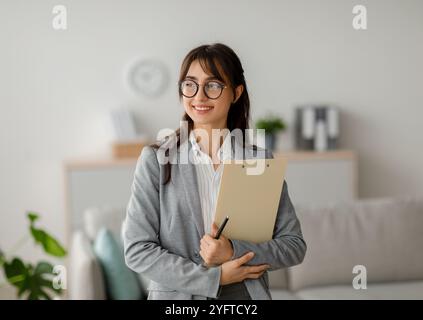Portrait de femme psychothérapeute arabe avec presse-papiers regardant de côté et souriant, debout au bureau moderne, espace libre Banque D'Images