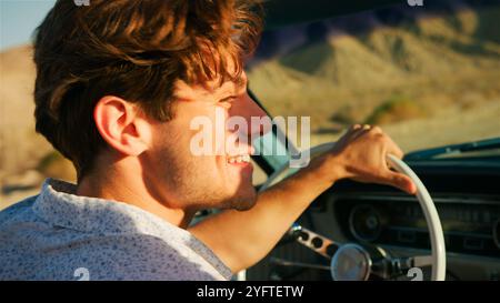 Gros plan d'un homme conduisant une voiture classique à toit ouvert sur la route à travers la campagne au coucher du soleil Banque D'Images