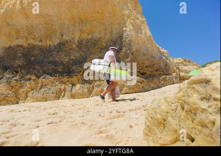 Un fournisseur dédié gravit les pentes sablonneuses de Praia de São Rafael tout en transportant des bolas de berlim traditionnels dans des glacières en polystyrène. Banque D'Images