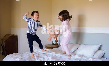 Enfants actifs s'amusant à sauter et jouer sur le lit des parents à la maison avec ours en peluche Banque D'Images