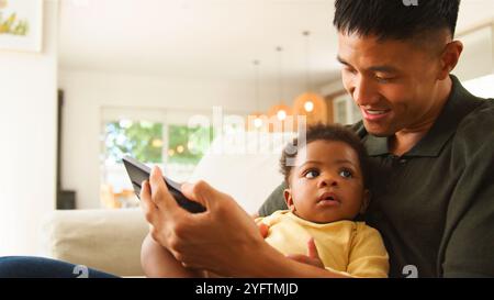 Un père est assis confortablement sur un canapé, tenant un appareil mobile tout en interagissant attentivement avec son bébé curieux. Le réglage intérieur chaud présente donc Banque D'Images