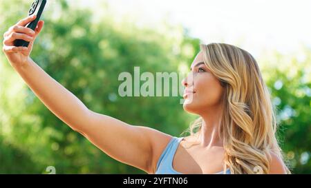 Une jeune femme se tient dehors, capturant un selfie avec son smartphone. Le feuillage vert vif en arrière-plan crée une atmosphère vibrante, highlig Banque D'Images