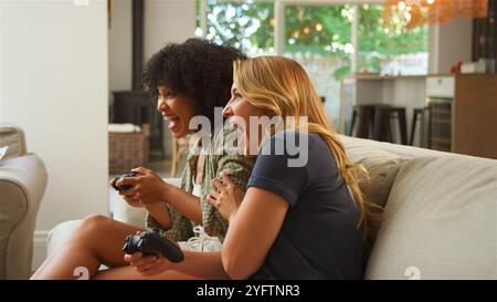 Deux amis assis sur un canapé confortable dans un salon confortable, immergé dans une session de jeu vidéo animée. Ils réagissent joyeusement au jeu, partageant Banque D'Images