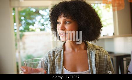 Une femme aux cheveux bouclés sourit tout en tenant un verre d'eau, rayonnant de contentement dans une cuisine ensoleillée. La verdure à l'extérieur complète le chaud at Banque D'Images