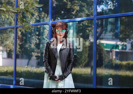 Une jeune femme heureuse dans une robe bleue et une veste en cuir noir, portant des lunettes bleues sur fond d'un mur de verre, regarde l'appareil photo. Banque D'Images