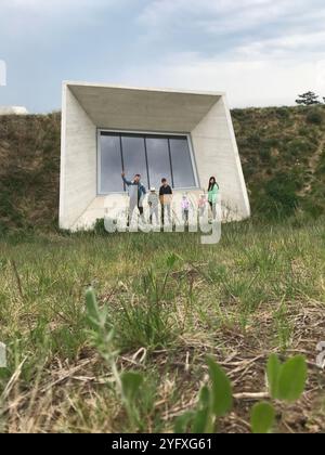 Une famille debout ensemble à l'extérieur d'un bâtiment moderne et géométrique avec de grandes fenêtres. Ils sont entourés d'herbe naturelle et de verdure, profitant d'une sortie Banque D'Images