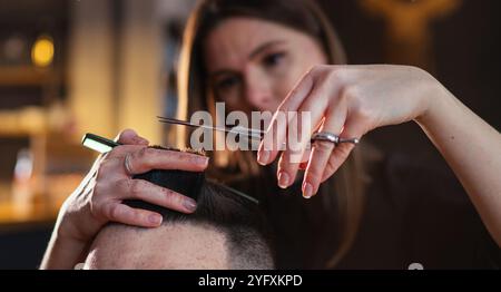 Jeune coiffeur mains féminines avec ciseaux et peigne coiffure coiffure image gros plan dans un salon de coiffure moderne de style noir à faible lumière. Service de soins capillaires Banque D'Images
