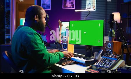 Musician fonctionne sur les touches de piano du contrôleur midi à côté de l'écran maquette, produisant de nouvelles chansons avec des instruments de musique et de l'équipement audio dans le home studio. L'artiste travaille avec un écran vert. Caméra B. Banque D'Images