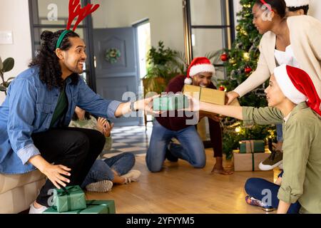 Porter des chapeaux de fête, des amis multiraciaux échangeant des cadeaux par arbre de noël, à la maison Banque D'Images