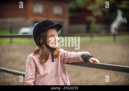 Petite fille dans un casque se tient sur un ranch, apprenant à monter à cheval. Portrait d'une enfant fille dans jockey un cheval, formation d'équitation. Petite fille a Banque D'Images