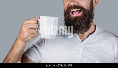 Gros plan portrait d'un homme barbu tenant une tasse et souriant. Homme heureux tenant une tasse de thé. Taille basse avec barbe tient mug. Un homme au visage heureux essaie de le faire Banque D'Images