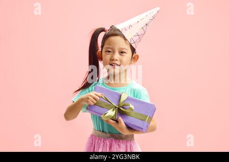 Charmante fille asiatique d'enfant autour de 5-6 ans dans le chapeau de fête, portant dans des vêtements de fête, tenant la boîte-cadeau sur fond isolé rose Banque D'Images