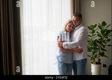 Couple heureux embrassant sur fond clair à la maison. Jolie femme souriant et embrassant son mari les yeux fermés. Couple doux en vêtements décontractés. Banque D'Images