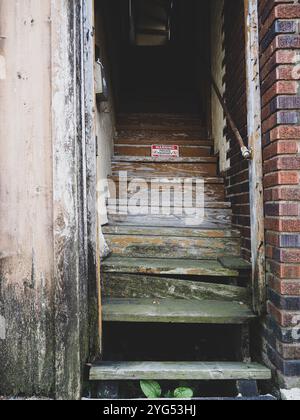 Des escaliers en bois altérés mènent à un couloir sombre dans un bâtiment abandonné avec des murs en pelage et un panneau d'avertissement Banque D'Images