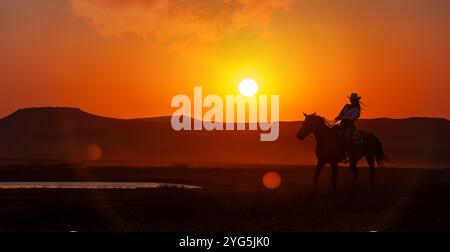 silhouette cow-girl dans le coucher du soleil chevauchant dans la prairie près d'un point d'eau Banque D'Images