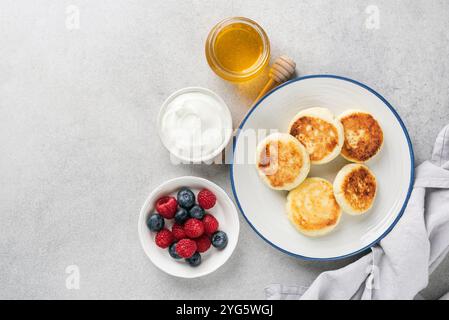 Syrniki, beignets au fromage cottage servis avec des baies fraîches, de la crème et du miel sur un fond de béton gris. Vue de dessus, espace de copie Banque D'Images