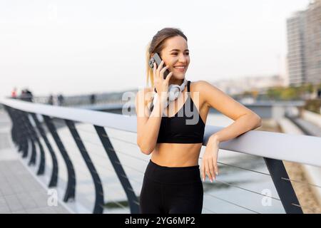 Une jogger s'arrête pour discuter sur son téléphone au bord de la rivière, portant un équipement d'entraînement et des écouteurs, embrassant le soleil chaud et l'atmosphère sereine arou Banque D'Images