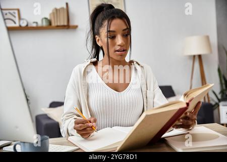 Une jeune femme concentrée étudie à distance, lisant un livre tout en prenant des notes à son bureau à domicile confortable. Banque D'Images