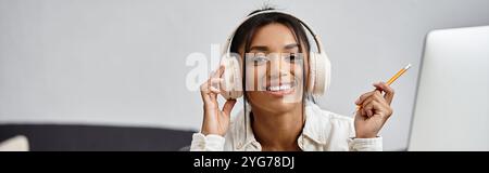 Une jeune femme joyeuse étudie à distance, souriante et portant des écouteurs dans son espace de travail. Banque D'Images