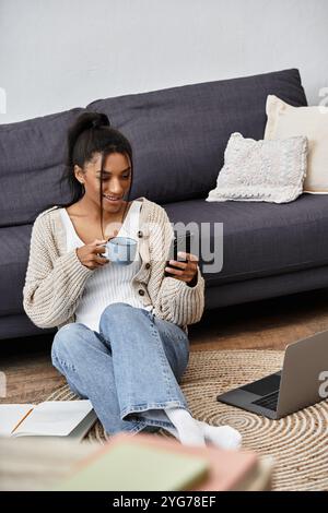Une femme joyeuse étudie à la maison, sirotant du thé et vérifiant son téléphone tout en étant assise confortablement. Banque D'Images