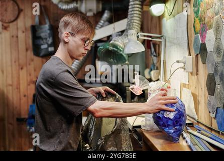 Jeune ouvrier masculin de l'atelier de recyclage en plastique prenant des bouchons de bouteilles emballés et les mettant dans un grand sac de cellophane Banque D'Images