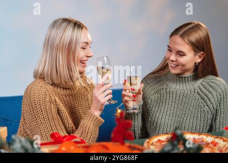 Deux femmes joyeuses appréciant le dîner de Noël, portant un toast avec du champagne et souriant, avec des cadeaux et des pizzas sur la table Banque D'Images