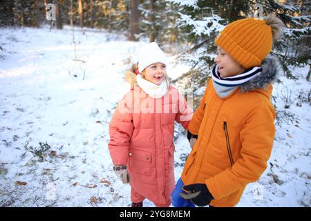 Un frère et une sœur marchent joyeusement ensemble à travers une forêt enneigée sereine, entourée de grands arbres recouverts de blanc. Banque D'Images