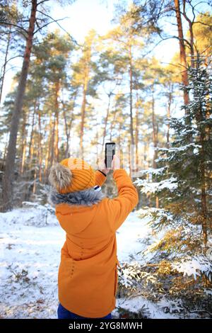 Un jeune homme se tient debout dans une forêt hivernale, capturant la beauté du paysage enneigé avec son smartphone. Banque D'Images