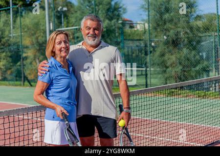 copyspace couple senior de joueurs de tennis partage un câlin joyeux, souriant à la caméra tout en tenant leurs raquettes sur le court du club Banque D'Images