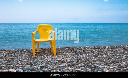 Une chaise en plastique jaune sur une plage de pierre en face de la mer calme contraste minimaliste Banque D'Images