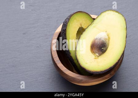 Deux moitiés d'avocat, l'une avec la fosse enlevée, sont nichées dans un bol en bois sur un fond gris. Banque D'Images