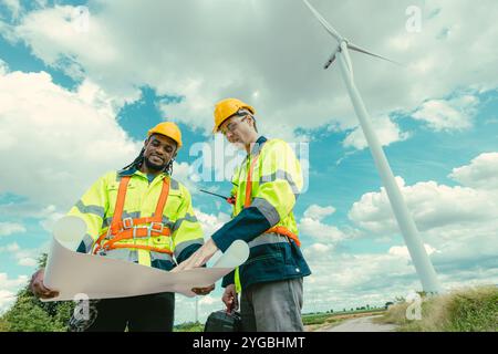 L'équipe d'ingénieurs mélange des ouvriers noirs et blancs masculins avec le plan d'étage à l'étude de travail sur le terrain de turbines éoliennes sur le terrain de ferme de générateur d'électricité éolienne en plein air. Banque D'Images