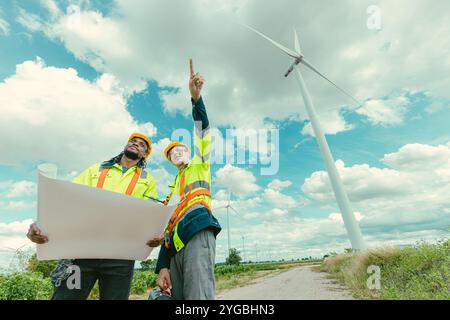 L'équipe d'ingénieurs mélange des ouvriers noirs et blancs masculins avec le plan d'étage à l'étude de travail sur le terrain de turbines éoliennes sur le terrain de ferme de générateur d'électricité éolienne en plein air. Banque D'Images