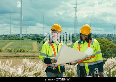 L'équipe d'ingénieurs mélange des ouvriers noirs et blancs masculins avec le plan d'étage à l'étude de travail sur le terrain de turbines éoliennes sur le terrain de ferme de générateur d'électricité éolienne en plein air. Banque D'Images