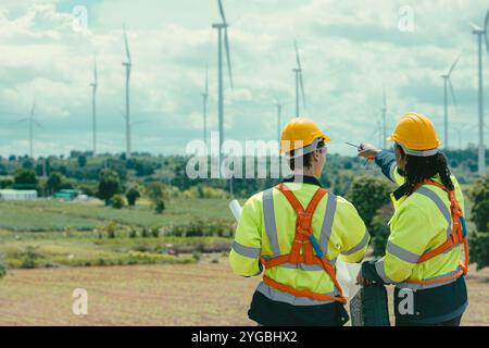 L'équipe d'ingénieurs mélange des ouvriers noirs et blancs masculins avec le plan d'étage à l'étude de travail sur le terrain de turbines éoliennes sur le terrain de ferme de générateur d'électricité éolienne en plein air. Banque D'Images