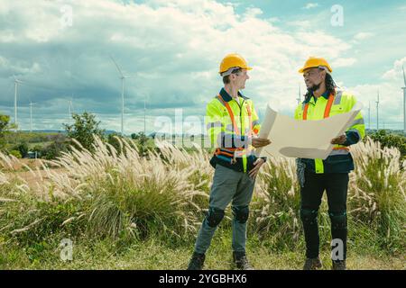 L'équipe d'ingénieurs mélange des ouvriers noirs et blancs masculins avec le plan d'étage à l'étude de travail sur le terrain de turbines éoliennes sur le terrain de ferme de générateur d'électricité éolienne en plein air. Banque D'Images