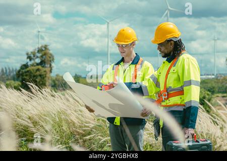 L'équipe d'ingénieurs mélange des ouvriers noirs et blancs masculins avec le plan d'étage à l'étude de travail sur le terrain de turbines éoliennes sur le terrain de ferme de générateur d'électricité éolienne en plein air. Banque D'Images