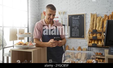 Jeune homme dans une boulangerie vérifiant son téléphone tout en se tenant debout derrière le comptoir avec du pain frais et des pâtisseries exposés Banque D'Images