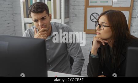 Homme et femme en tenue d'affaires travaillant ensemble et regardant soigneusement un écran d'ordinateur à l'intérieur dans une pièce bien éclairée avec un tableau d'affichage en th Banque D'Images