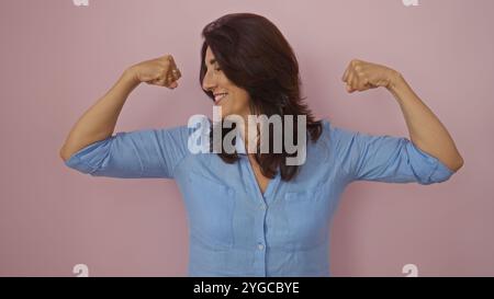 Femme hispanique d'âge moyen en chemise bleue fléchissant les muscles en toute confiance sur fond rose Banque D'Images