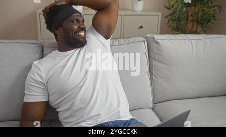 Un beau jeune homme afro-américain avec une barbe est confortablement assis dans un salon bien meublé, souriant et à l'air détendu. Banque D'Images