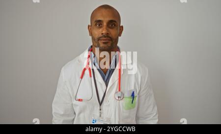 Beau jeune homme afro-américain dans un manteau blanc avec un stéthoscope et un badge d'identification, debout avec confiance contre un dos blanc isolé Banque D'Images