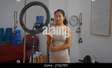 Femme posant dans une salle de sport avec un équipement d'entraînement et un smartphone sur un trépied, démontrant des exercices de fitness dans un cadre intérieur, créant du contenu pour Banque D'Images